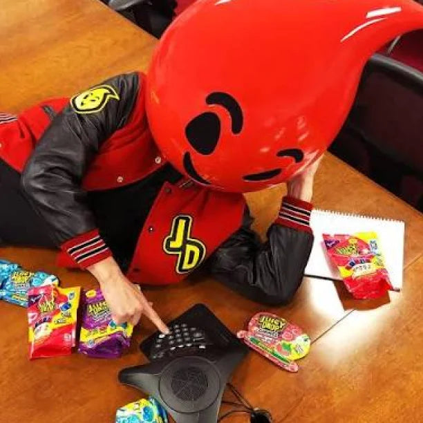 A person wearing a red and black varsity jacket and a large red teardrop-shaped mascot head sits on a wooden table, pointing at a black conference phone. Several colorful candy bags are scattered around the table.
