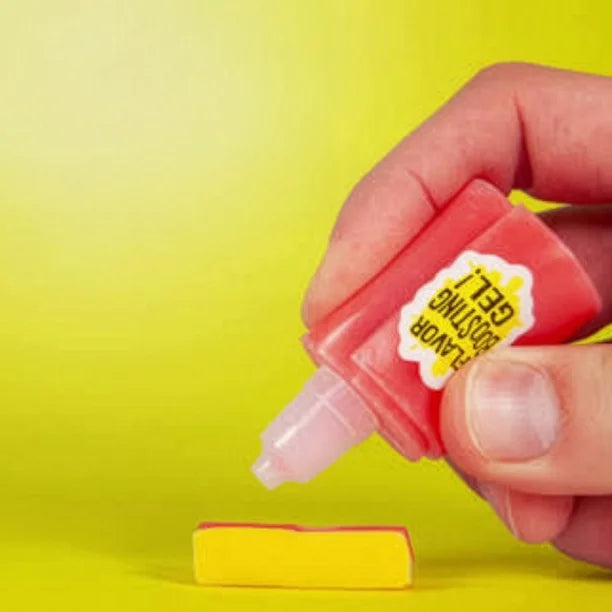A hand holding a small pink bottle of Flavour Boosting Gel, which is shown dispensing a pink gel onto a yellow rectangular piece against a bright yellow background.