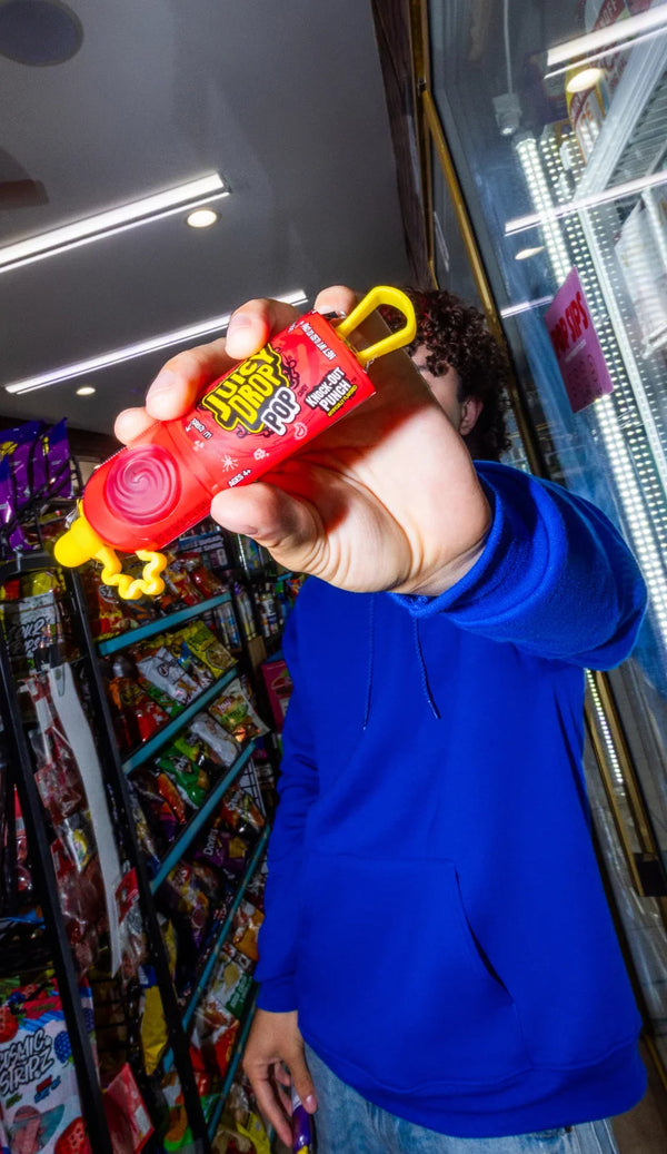 A person in a blue hoodie holds up a small red and yellow novelty toy bottle with a yellow loop handle in a retail store aisle.